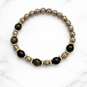 Peut inclure: Un bracelet de perles avec un mélange de textures et de couleurs. Le bracelet comprend des perles rondes vert foncé et noires, entrecoupées de perles texturées de couleur or. Le bracelet est posé sur un fond de marbre blanc.