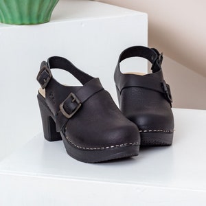 May include: A pair of black leather clogs with a high heel and buckle details. The clogs have a platform sole with white stitching and a strap around the ankle. A green vase is in the background.