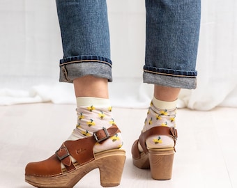 May include: Brown leather clogs with a buckle strap and a wooden heel. The clogs are worn with white socks that have a yellow floral pattern.