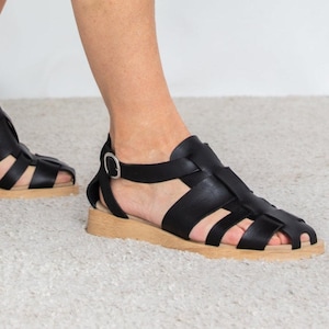 May include: Black leather sandals with a cage-like design. The sandals have a light brown sole and a buckle closure around the ankle. The sandals are on a light-colored carpet.