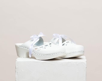 May include: White leather clogs with a white ribbon tied around the ankle. The clogs have a cutout design and a chunky heel.
