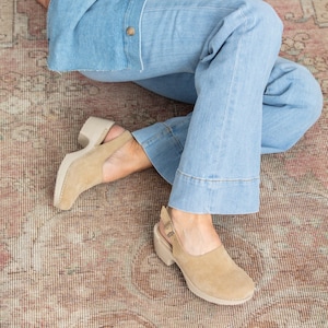 May include: A pair of light brown suede clogs with a slingback strap and a chunky heel. The clogs are worn by a person wearing light blue jeans.