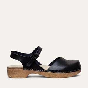 May include: Black leather clog sandal with a wooden sole and an adjustable ankle strap. The sandal features a closed toe design and a buckle closure. The wooden sole is approximately 2 inches high.