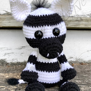 Puede incluir: Un animal de peluche de cebra de crochet a rayas blancas y negras con grandes ojos negros. La cebra está sentada sobre una superficie de madera.
