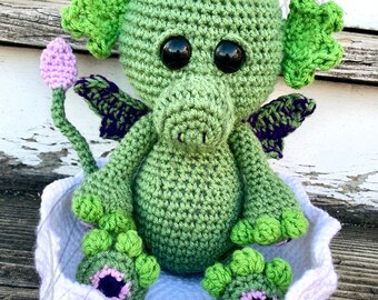 May include: A green crocheted dragon with purple and pink accents sits in a white crocheted nest. The dragon has large black eyes and a long tail.