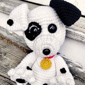 Puede incluir: Un perro dálmata de crochet blanco con manchas negras, ojos grandes negros y una oreja negra. El perro lleva un collar rojo con una etiqueta circular dorada.