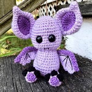 May include: A crocheted purple bat with black eyes and wings. The bat has a small mouth and is sitting on a wooden surface.
