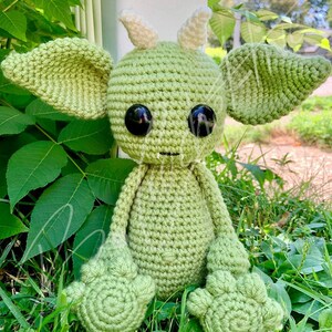 May include: A green crocheted amigurumi toy with large eyes and pointed ears. The toy is sitting on a bed of green grass.