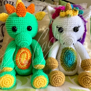 May include: A green crocheted dragon and a white crocheted unicorn with rainbow details. Both toys have large black eyes and are sitting on a bed.