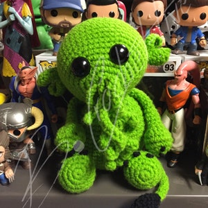 May include: Green crocheted Cthulhu plush toy with black eyes and a black tail. The toy is sitting on a shelf with other figurines.