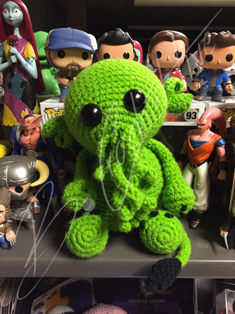 May include: Green crocheted Cthulhu plush toy with black eyes and a black tail. The toy is sitting on a shelf with other figurines.