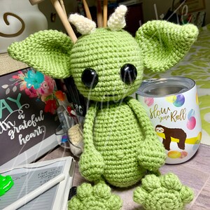 May include: A green crocheted amigurumi monster with large black eyes and white horns. The monster is sitting on a table with a white and green tumbler with the text "Slow your Roll" and a sloth graphic.