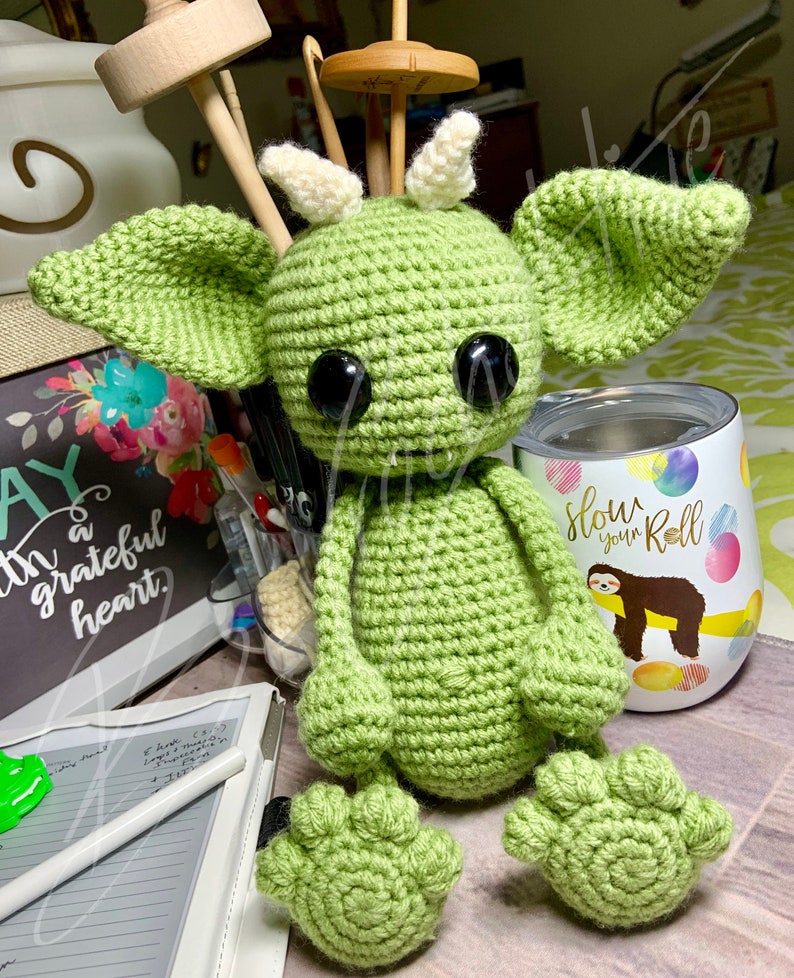 May include: A green crocheted amigurumi monster with large black eyes and white horns. The monster is sitting on a table with a white and green tumbler with the text "Slow your Roll" and a sloth graphic.