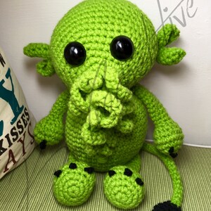 May include: Green crocheted plush toy with black eyes and a tail. The toy has a tentacle-like mouth and black claws.