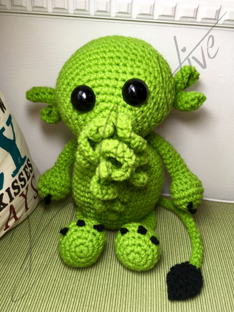 May include: Green crocheted plush toy with black eyes and a tail. The toy has a tentacle-like mouth and black claws.