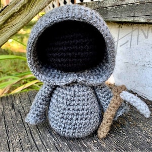 May include: A gray crocheted amigurumi figure of the Grim Reaper with a black hooded face and a brown and gray scythe.