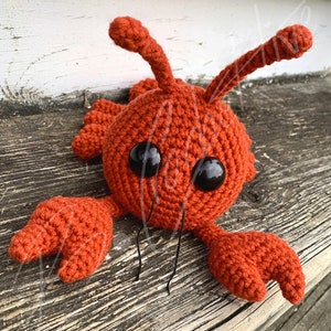 May include: A crocheted orange crab toy with black eyes and antennae. The crab is lying on a wooden surface.
