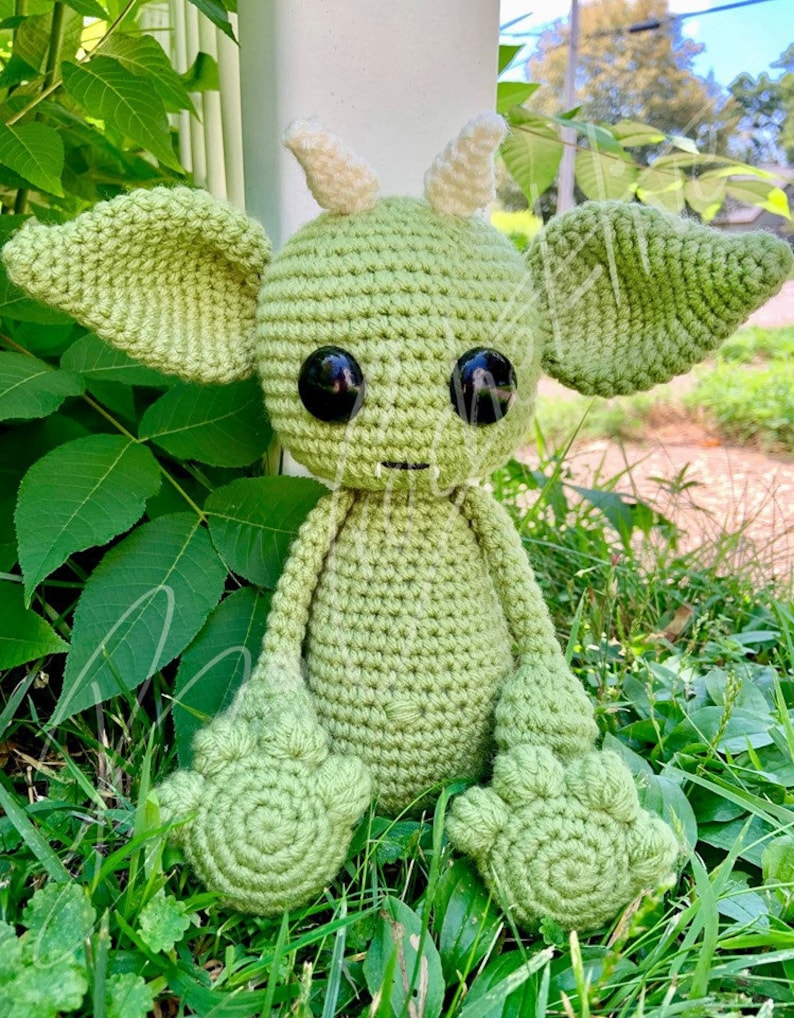 May include: A green crocheted amigurumi toy with large eyes and pointed ears. The toy is sitting on a bed of green grass.