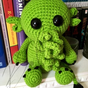 May include: Green crocheted plush toy with black eyes and a tentacle-like tail. The toy is sitting on a shelf with books behind it.
