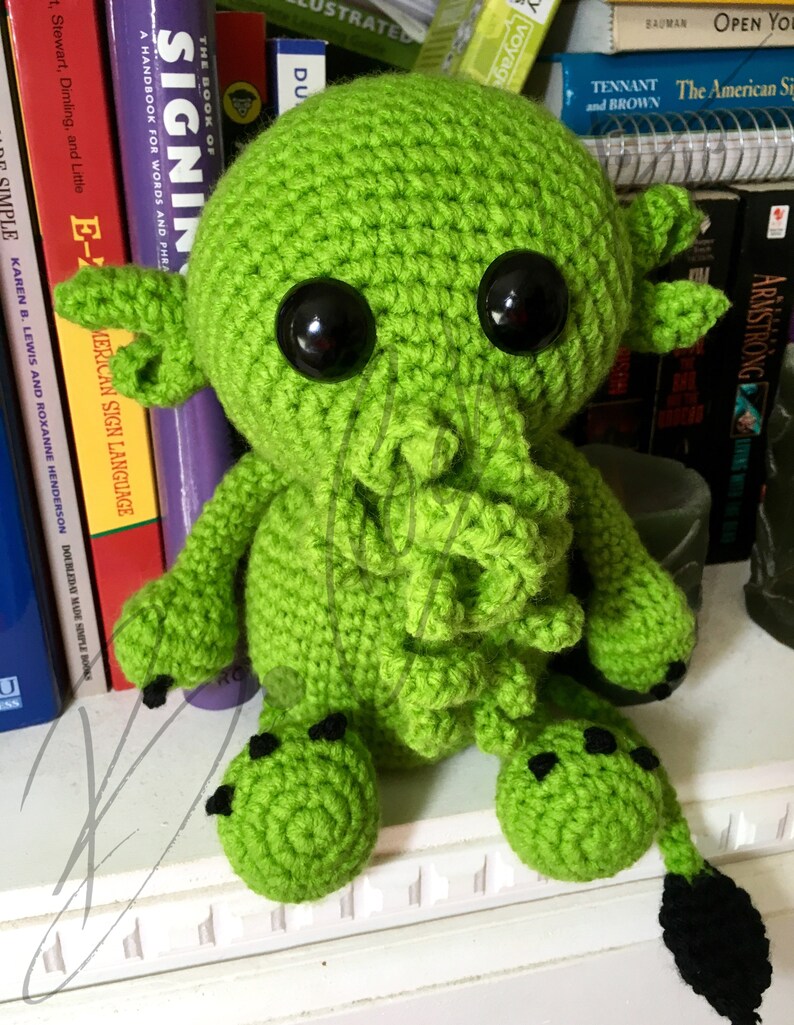 May include: Green crocheted plush toy with black eyes and a tentacle-like tail. The toy is sitting on a shelf with books behind it.