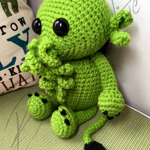 May include: Green crocheted plush toy with black eyes and a long tail. The toy has a tentacle-like head and small feet.