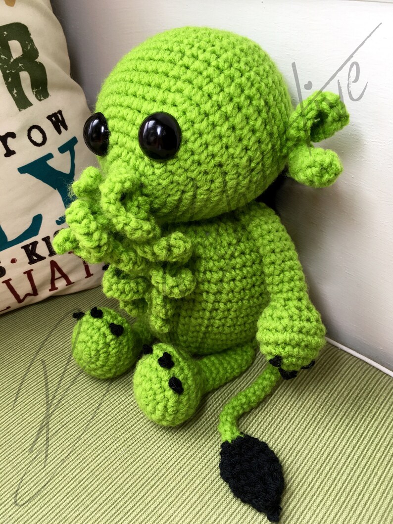 May include: Green crocheted plush toy with black eyes and a long tail. The toy has a tentacle-like head and small feet.