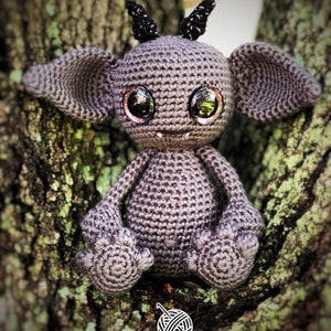 May include: A crocheted grey monster with black horns and large pink eyes. The monster is sitting on a tree trunk with green moss.