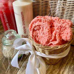 May include: A wicker basket filled with a pink crocheted washcloth, tied with a white satin ribbon. Two bottles of body wash with the label "apple blossom" are in the background.