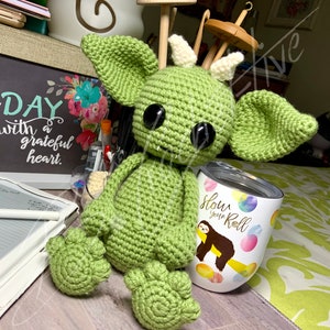 May include: Green crocheted amigurumi monster with large black eyes and white horns. The monster is sitting on a surface with a white tumbler that says "Slow Your Roll" with a cartoon sloth on it.