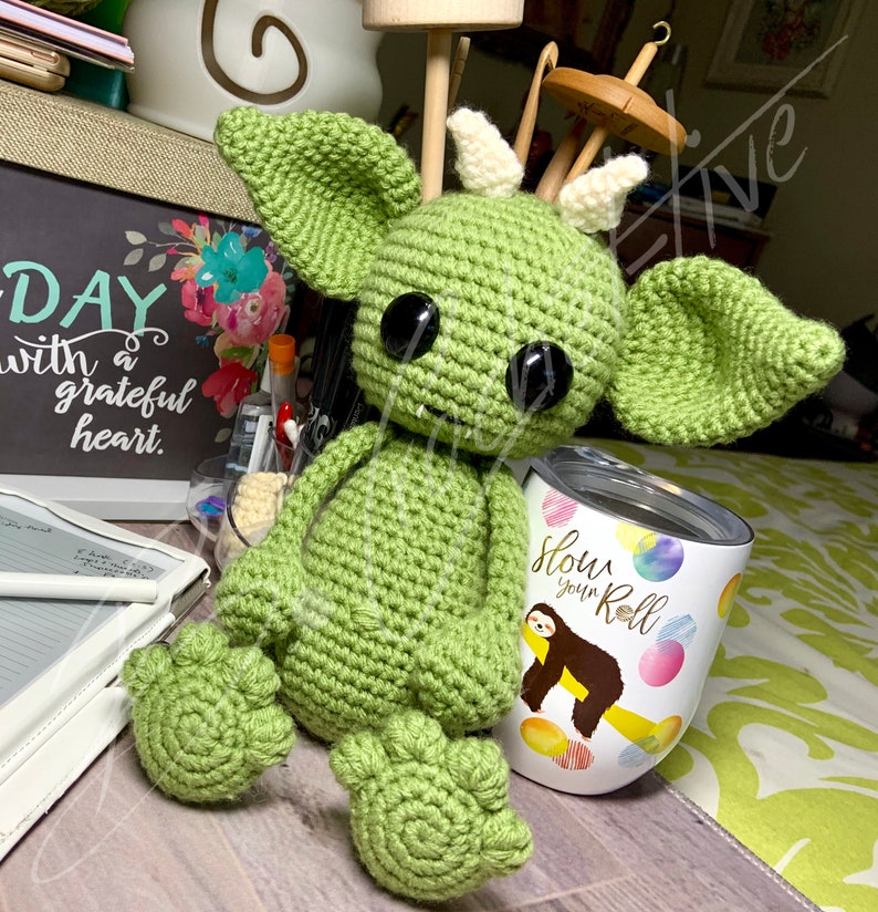 May include: Green crocheted amigurumi monster with large black eyes and white horns. The monster is sitting on a surface with a white tumbler that says "Slow Your Roll" with a cartoon sloth on it.