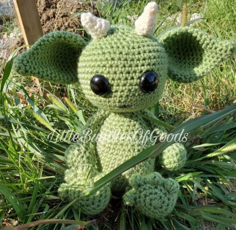May include: A green crocheted plush toy with large black eyes and white horns. The toy is sitting in a bed of green grass.