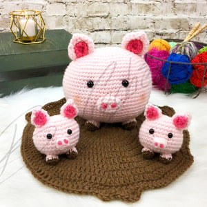 May include: Three crocheted pink pig plushies. The largest pig is sitting on a brown crocheted mat. The two smaller pigs are sitting on either side of the larger pig.
