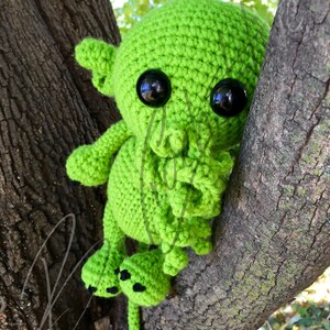 May include: A green crocheted creature with black eyes and a long tail, sitting on a tree branch.