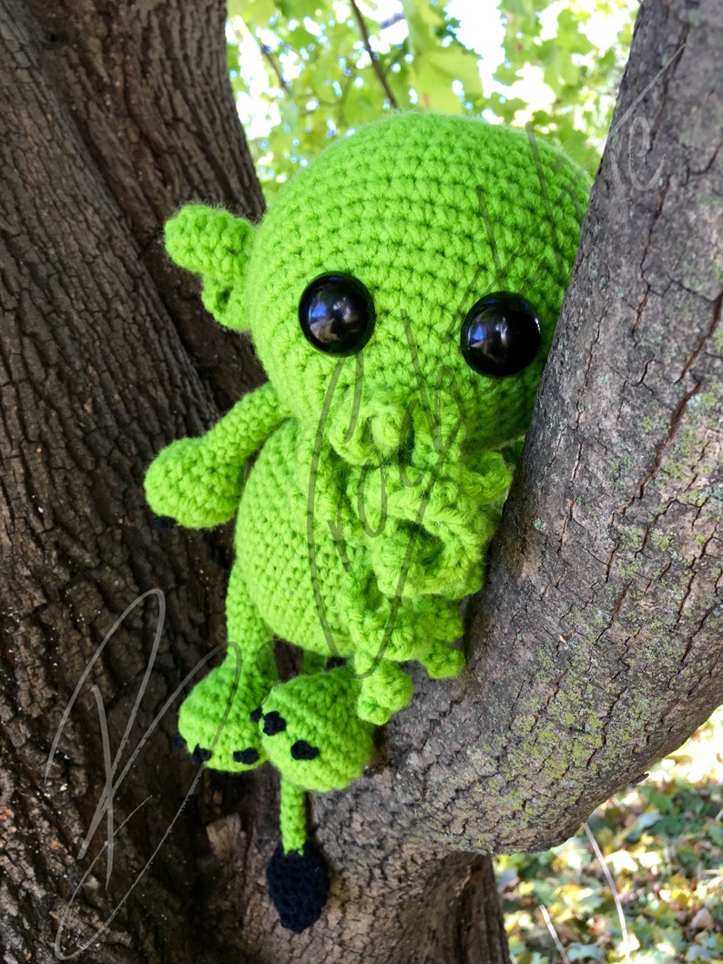 May include: A green crocheted creature with black eyes and a long tail, sitting on a tree branch.