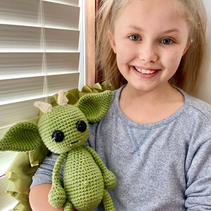 May include: A young person is holding a green crocheted plush toy that resembles a goblin or a monster. The toy has large eyes, a toothy grin, and pointy ears.