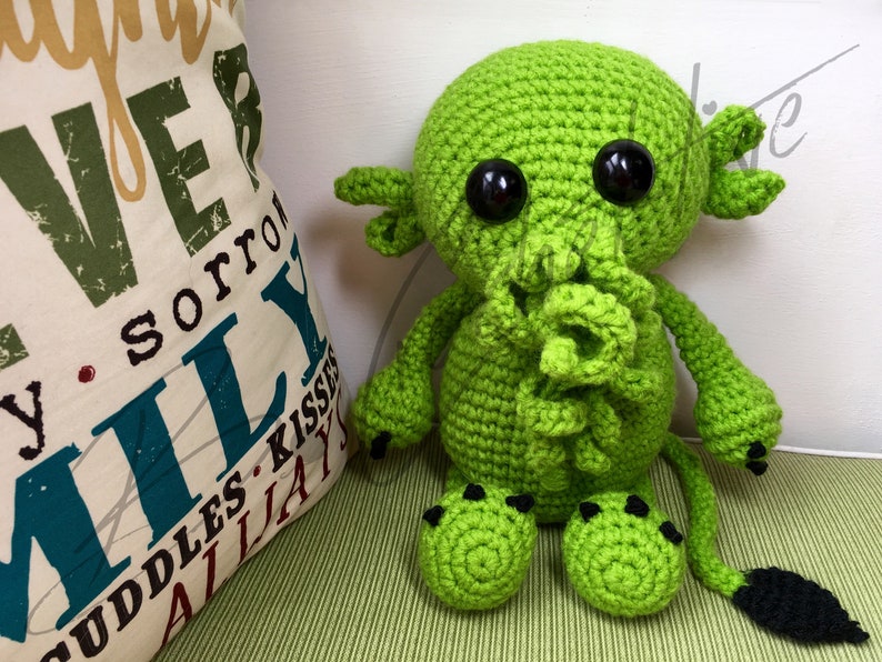 May include: Green crocheted plush toy with black eyes and a black tail. The toy is sitting on a green and white striped surface.