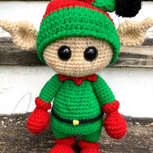 Op de afbeelding: Een gehaakte elfpop met een groene muts, rode laarzen en groene en rode kleding. De elf heeft grote bruine ogen en een beige gezicht.