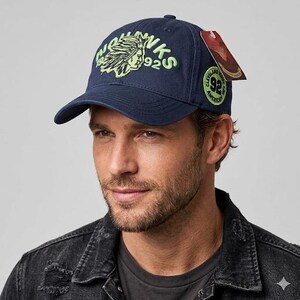 Op de afbeelding: Marineblauwe baseballcap met een gebogen klep. De cap heeft een neon groen en wit geborduurd logo met de tekst "Hawks 92" en een ontwerp van een Indiaanse chef. Een cirkelvormige patch met tekst staat aan de zijkant.