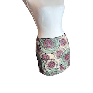 Può includere: Mini gonna beige con un motivo circolare ripetuto in rosa, verde e nero. La gonna, esposta su un manichino, ha una fascia in vita nera. Il motivo ricorda fiori o mandala stilizzati.