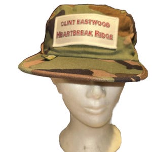 Puede incluir: Sombrero de pescador de camuflaje con un parche rectangular que dice "CLINT EASTWOOD HEARTBREAK RIDGE" en letras rojas y blancas. El sombrero es en tonos verdes, marrones y beiges.