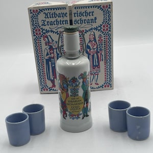 Könnte beinhalten: Eine weiße Keramikflasche mit Bügelverschluss, beschriftet mit "200 ml Bavarian Monastery Gold Liqueur", und vier kleine blaue Schnapsgläser. Die Flasche ist mit farbenfrohen Figuren und floralen Designs verziert und wird von einer dekorativen Schachtel begleitet.