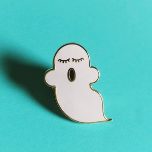 Puede incluir: Pin de esmalte blanco de un fantasma con los ojos cerrados y la boca abierta. El fantasma está rodeado de oro.
