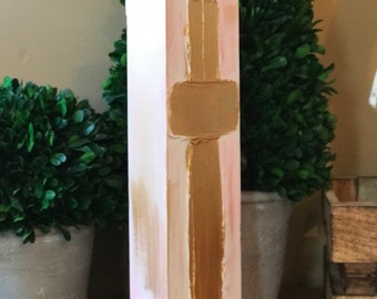 May include: A vertical wooden sign with a pink and white background and a gold cross painted on it.