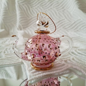 Puede incluir: Adorno de tetera de cristal rosa con detalles dorados. La tetera tiene un asa, un pico y una tapa transparentes, y está adornada con pequeños topes dorados. Se adjunta una cuerda dorada para colgar.