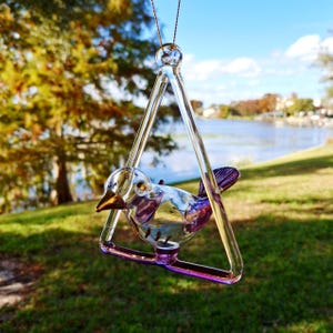 Puede incluir: Un adorno de pájaro de cristal transparente con cola y pico morados, suspendido dentro de un marco triangular. El adorno cuelga de un hilo dorado fino, con un fondo borroso de un lago y árboles.