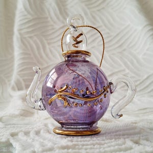 May include: A decorative glass teapot with a round body and a clear handle and spout. The teapot is primarily purple with gold accents and a clear glass lid. The teapot is sitting on a white surface.