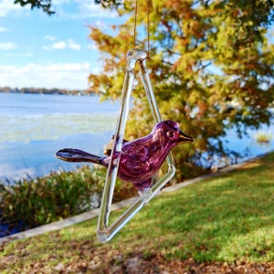 Puede incluir: Un adorno decorativo de pájaro de cristal púrpura, suspendido dentro de un marco triangular transparente, colgado de una cadena dorada. El pájaro tiene detalles dorados. El fondo muestra un lago, árboles y un cielo azul.