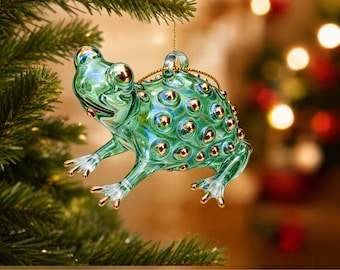 May include: A translucent green glass frog ornament featuring a textured back with raised gold-colored dots. The frog has gold-colored accents on its eyes and toes, and includes a small loop at the top for hanging with a thin gold string.