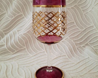 May include: A clear wine glass with a gold rim and a gold diamond pattern. The top and base of the glass are a deep purple color with a gold rim. The glass is on a white textured surface.