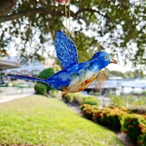 May include: A bluebird ornament with a gold hanging string. The bird has a blue body with white and brown accents, a textured blue wing, and a gold beak. The background is a blurred outdoor scene with green grass and trees.